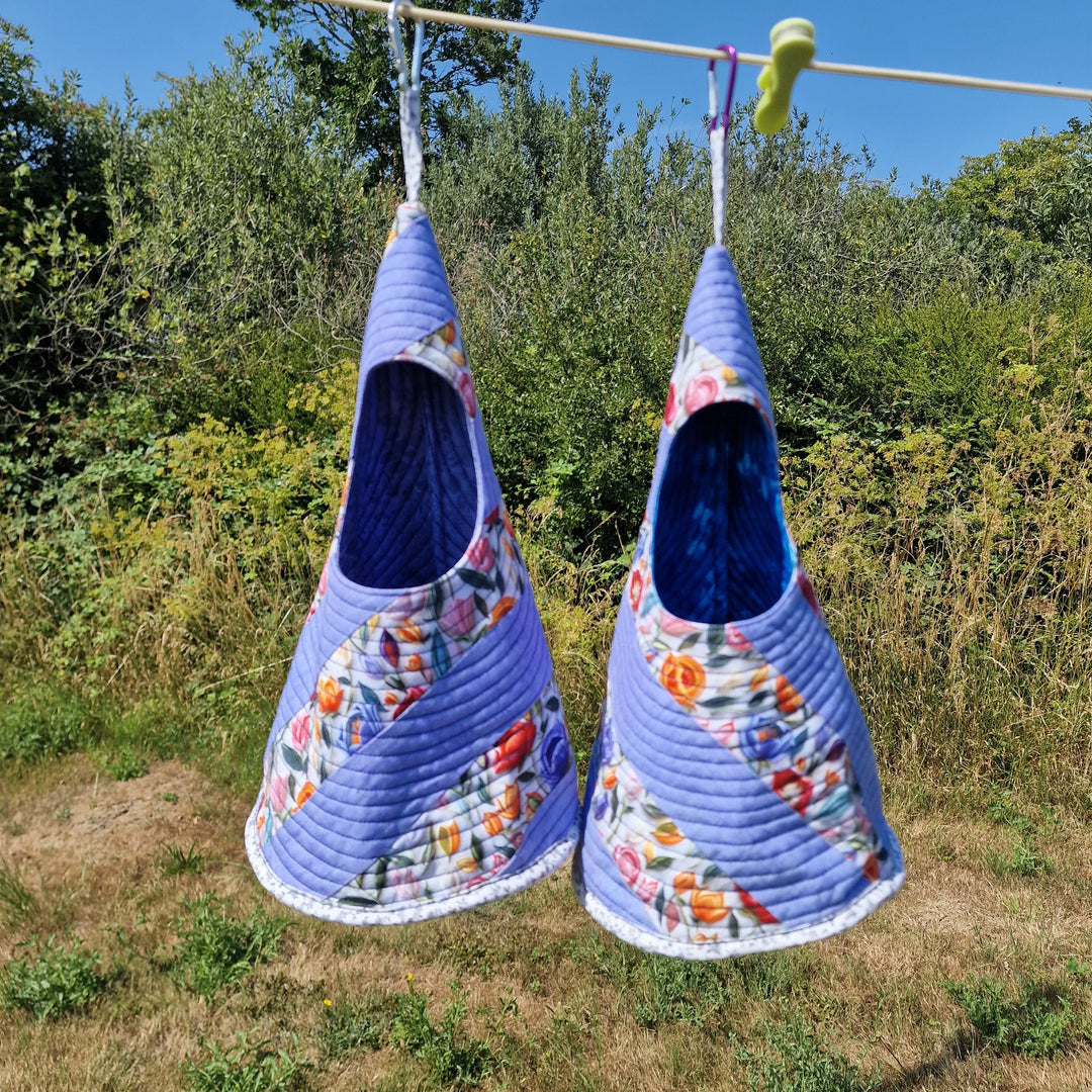 Two quilted fabric hanging peg bags in a lavender-blue floral print with a dark blue interior, hanging from a clothesline outdoors on a sunny day with green trees and grass in the background.