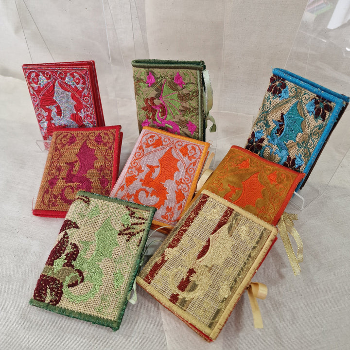 A group of small embroidered fabric Needlecases displayed on a light fabric surface with clear acrylic stands. The set includes multiple colours and patterns, such as red, green, orange, blue, and beige with detailed floral and animal embroidery.