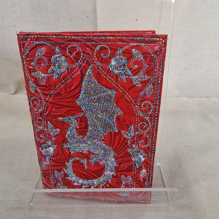 A red  embossed vegan leather Needlebook with intricate silver glitter dragon and scroll designs on the cover, displayed on a clear acrylic stand against a neutral beige fabric background.