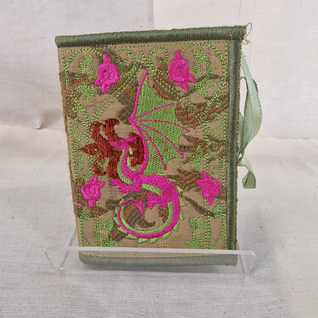 An embroidered fabric Needlebook cover with a green border, featuring a pink dragon design and pink flowers on a beige background, displayed on a clear stand against a light fabric backdrop.