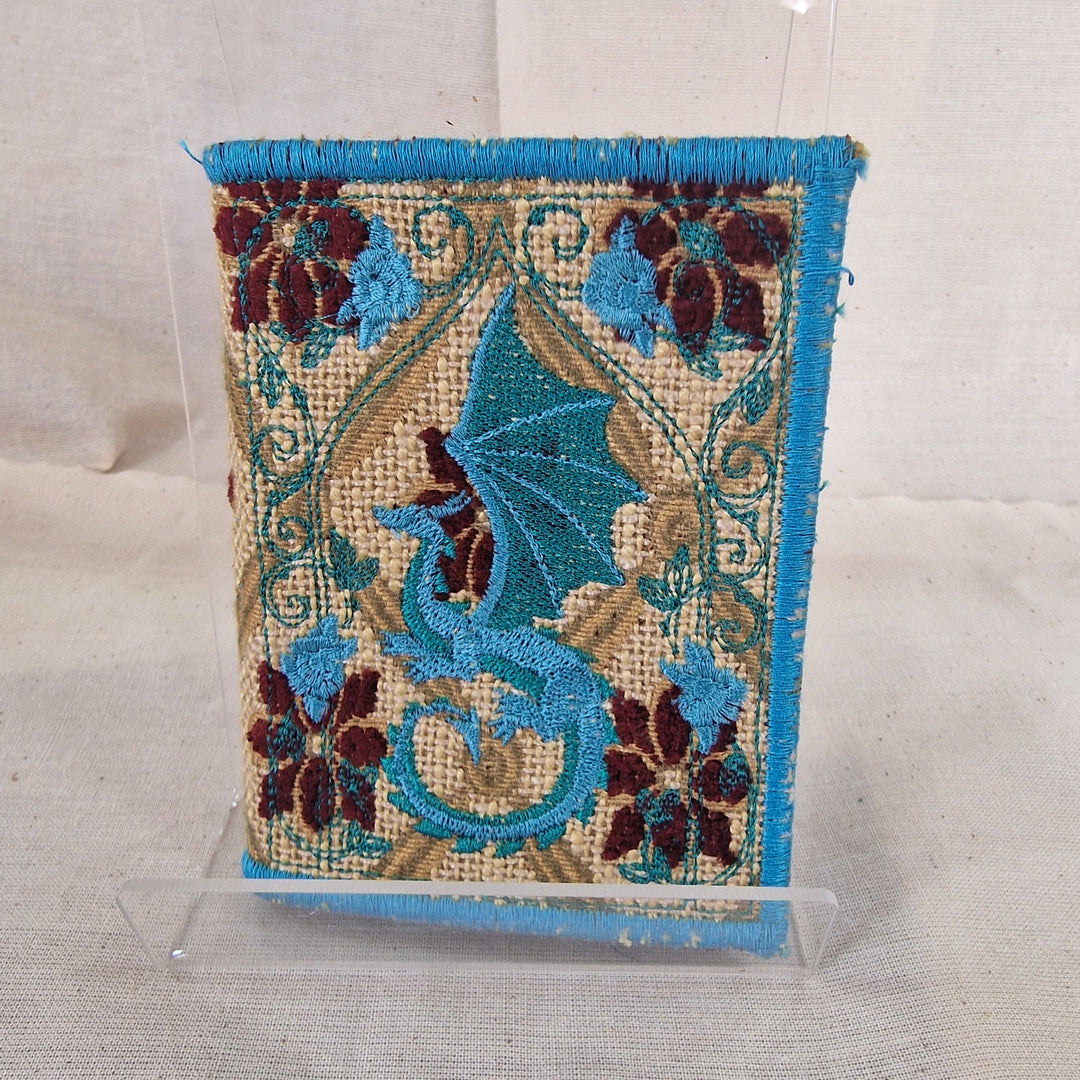 A Needlebook embroidered textile piece with a blue dragon motif, floral border details, and a turquoise fringe, displayed upright on a clear acrylic stand against a light fabric backdrop.