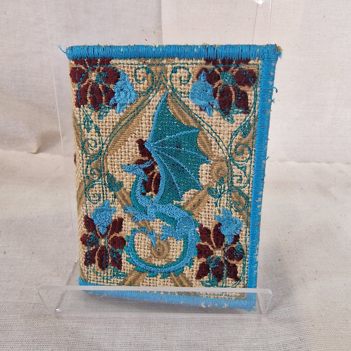A Needlebook embroidered textile piece with a blue dragon motif, floral border details, and a turquoise fringe, displayed upright on a clear acrylic stand against a light fabric backdrop.