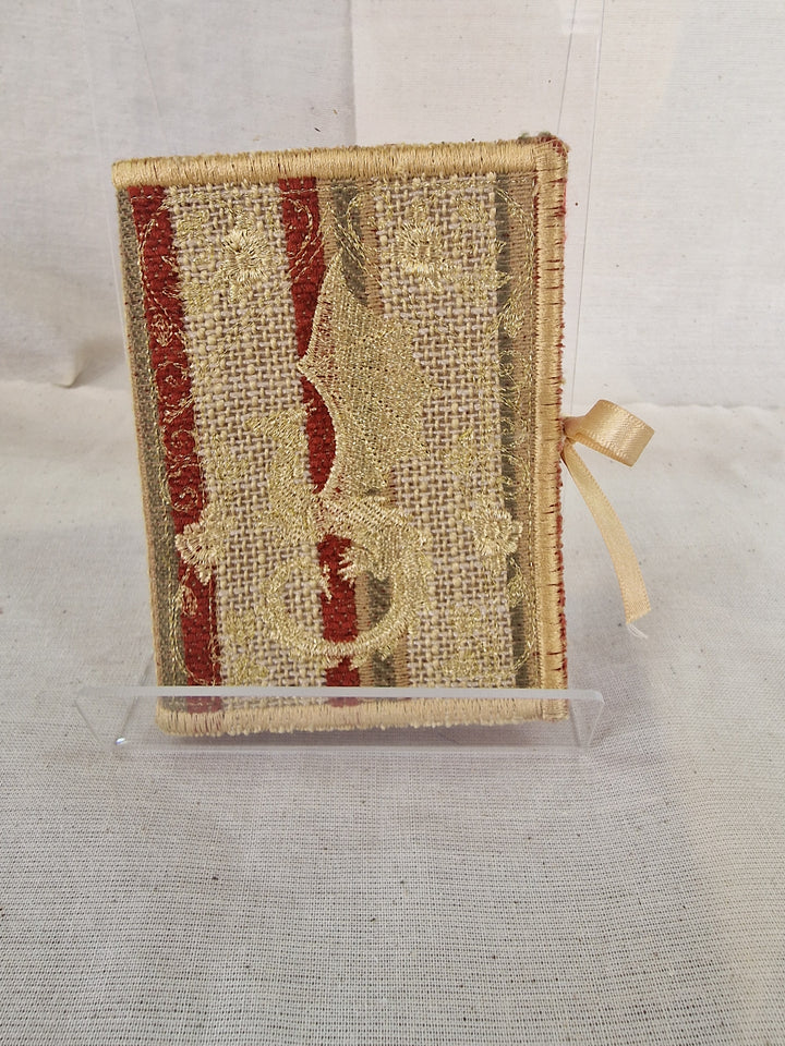 A small decorative Needlebook with a textured beige burlap-style cover, featuring red vertical stripes, a gold dragon motif, and a cream ribbon bookmark, displayed on a clear stand against a light fabric backdrop.