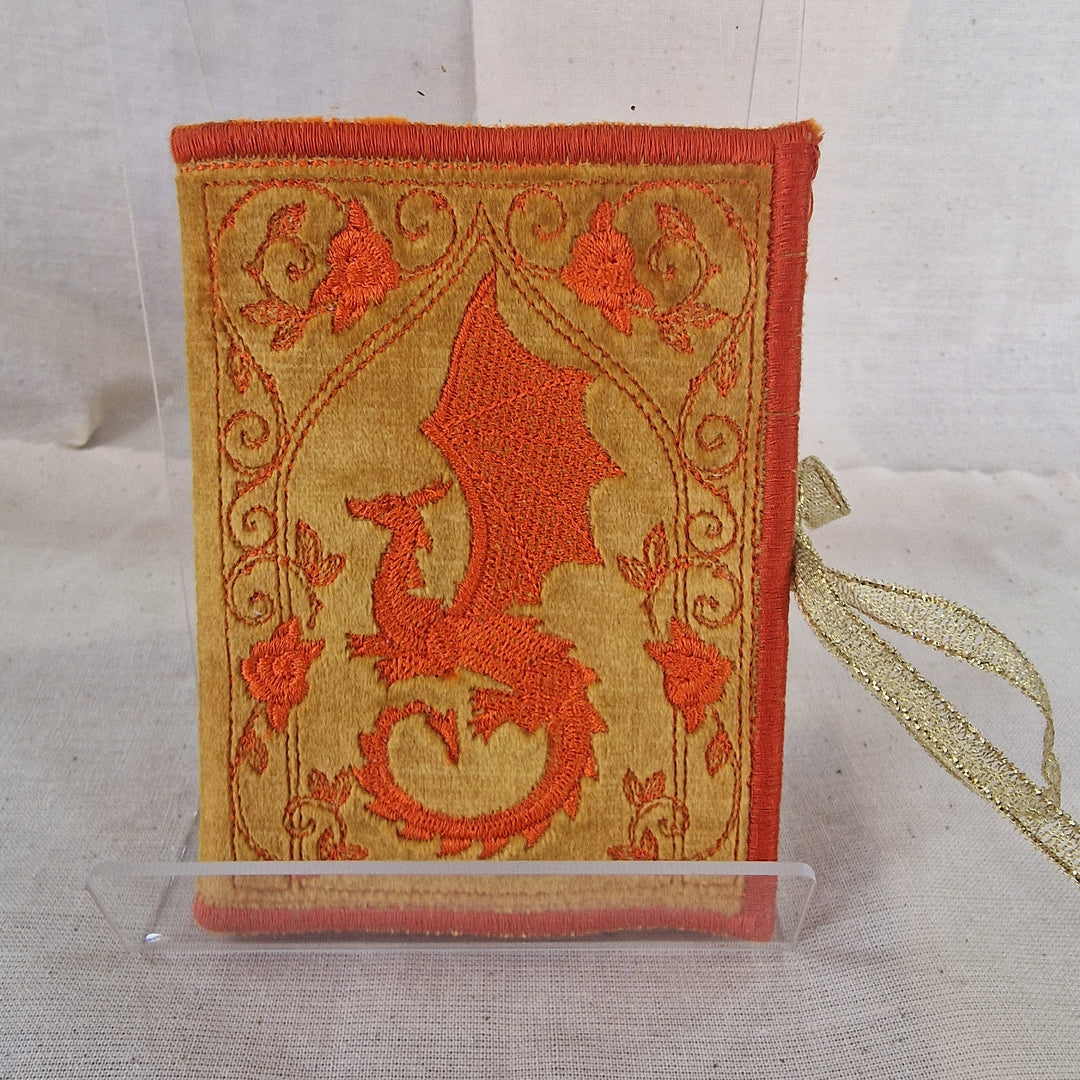 A small decorative fabric Needlebook with an embroidered dragon design in orange-red on a tan background, displayed upright in a clear stand against a light fabric backdrop.