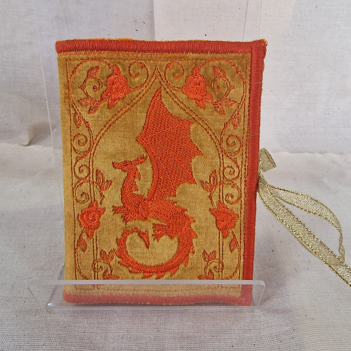 A small decorative fabric Needlebook with an embroidered dragon design in orange-red on a tan background, displayed upright in a clear stand against a light fabric backdrop.