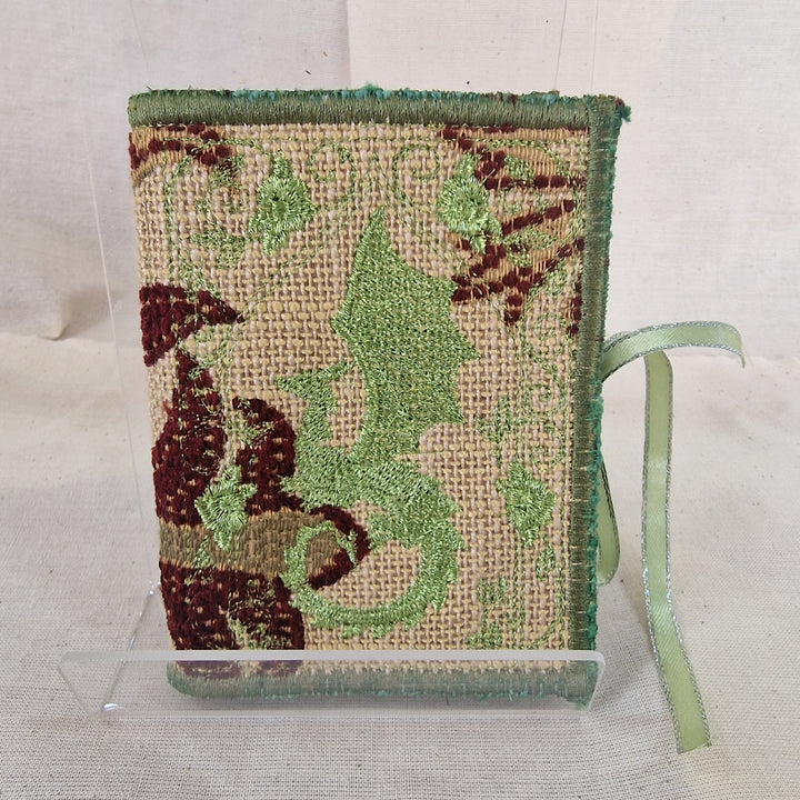 A small woven fabric needlebook with a green Dragon and ribbon  displayed on a clear acrylic stand against a neutral fabric background.
