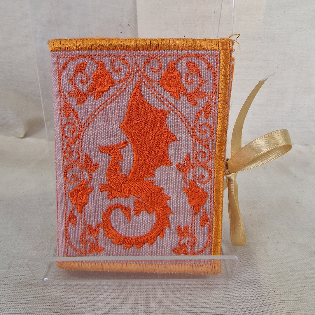 An embroidered fabric Needlebook featuring a prominent orange dragon design on a light pink background, finished with an orange border and a gold ribbon bookmark, displayed in a clear acrylic stand.