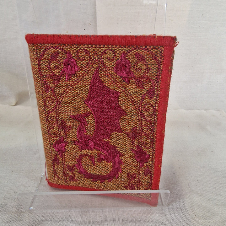 A small rectangular embroidered fabric Needlebook cover with a red border and a central dragon motif, displayed upright on a clear acrylic stand against a light fabric backdrop.