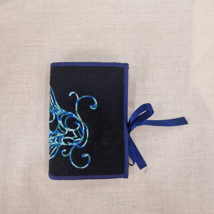 A small black fabric Needlecase  with blue embroidered fish details on the cover and a blue ribbon bookmark, laid flat on a light beige linen background.