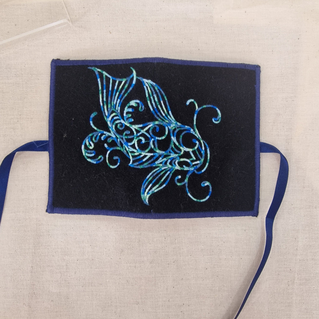 A square black fabric Needlecase with a blue embroidered fish design, finished with a blue border and matching blue ribbon ties, laid flat on a beige linen background.