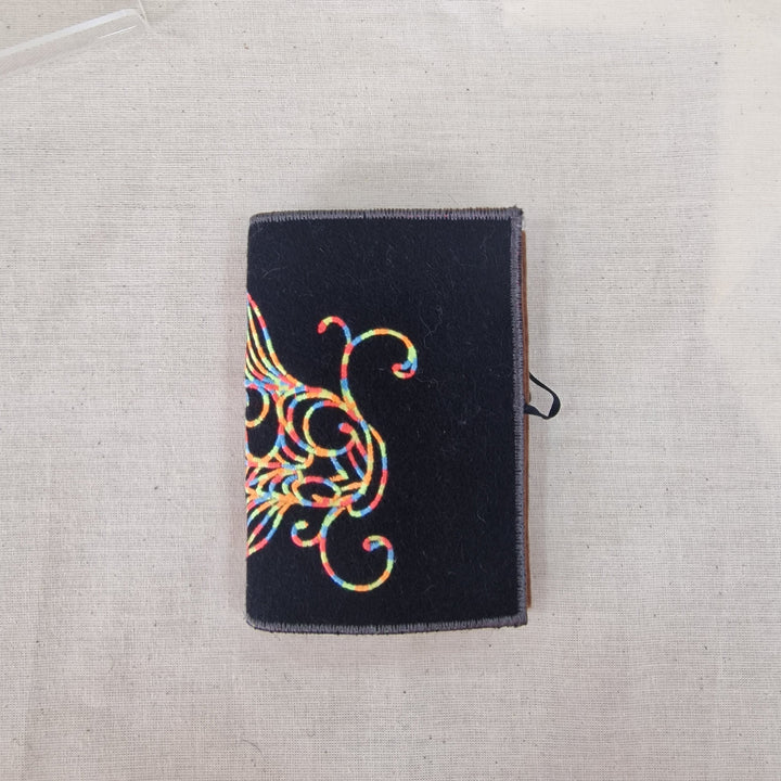 A black fabric Needlebook with a colourful embroidered fish design on the front, resting on a light beige linen fabric background.