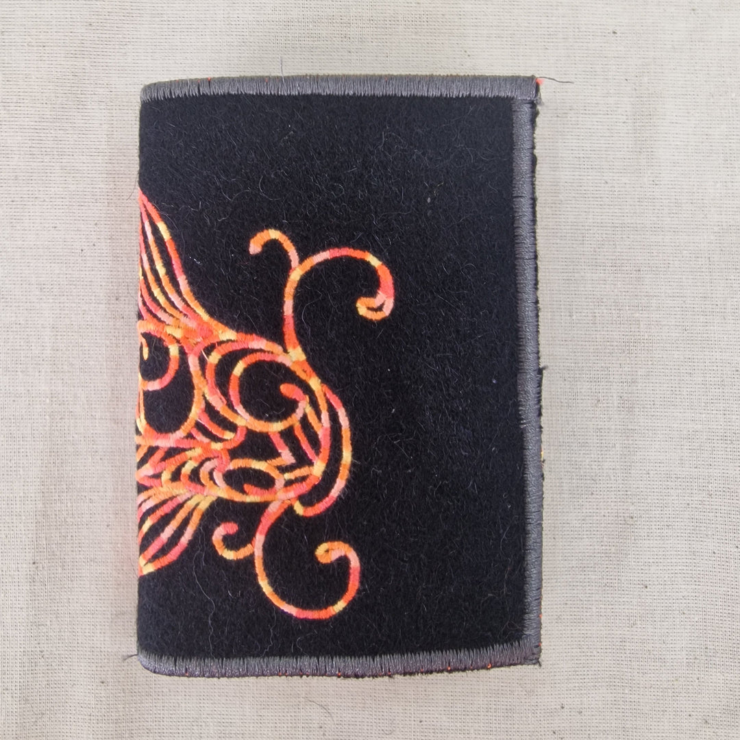 A rectangular black fabric Needlecase  with a gray stitched border and an orange embroidered fish shown on a light beige textured fabric background.