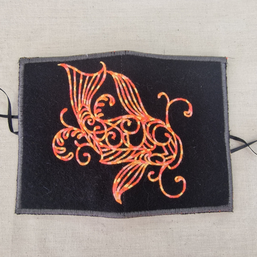 A square black fabric Needlecase with a gray border, featuring an intricate orange and red embroidered fish design, laid flat on a beige linen background.