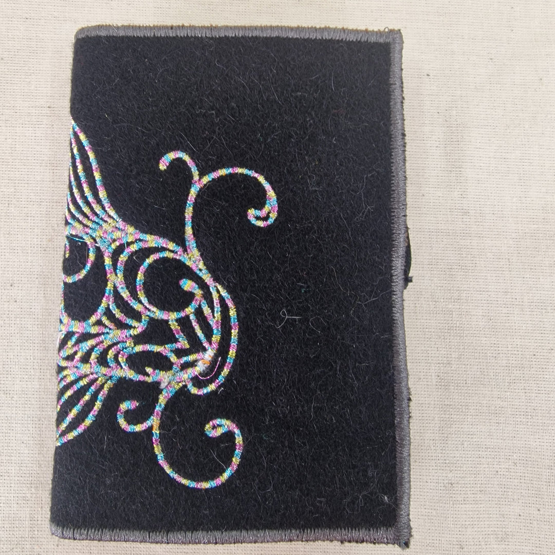 A rectangular black fabric needlecase with a gray stitched border and a silver embroidered fish  design on the left side, shown on a light beige textured fabric background.