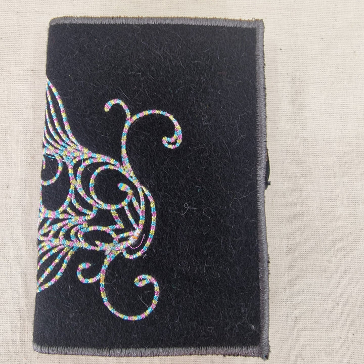 A rectangular black fabric needlecase with a gray stitched border and a silver embroidered fish  design on the left side, shown on a light beige textured fabric background.