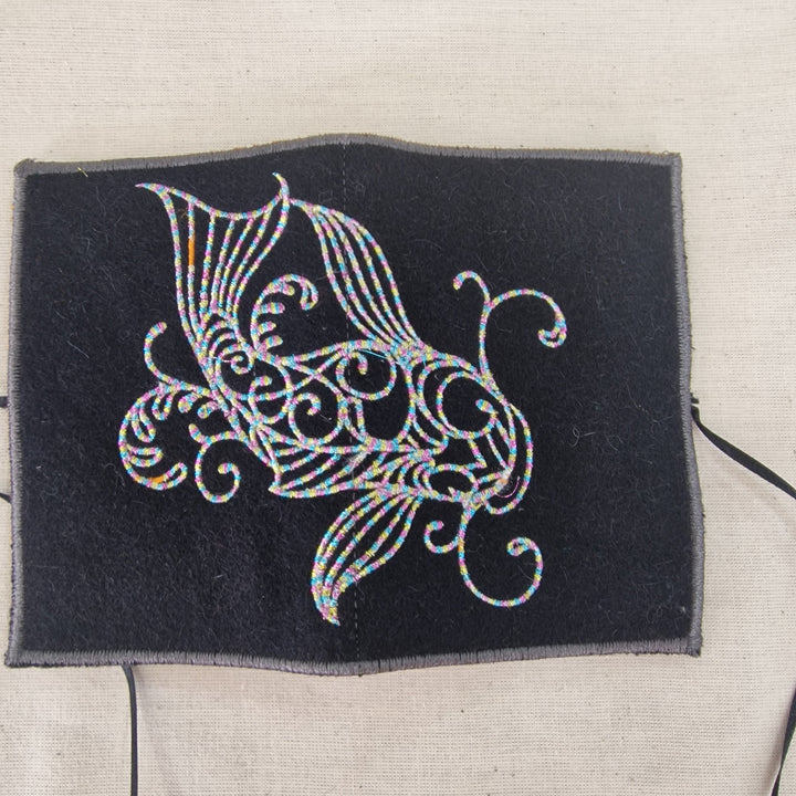 A square black fabric needlecase  with a white embroidered fish design, displayed on a light beige linen background.