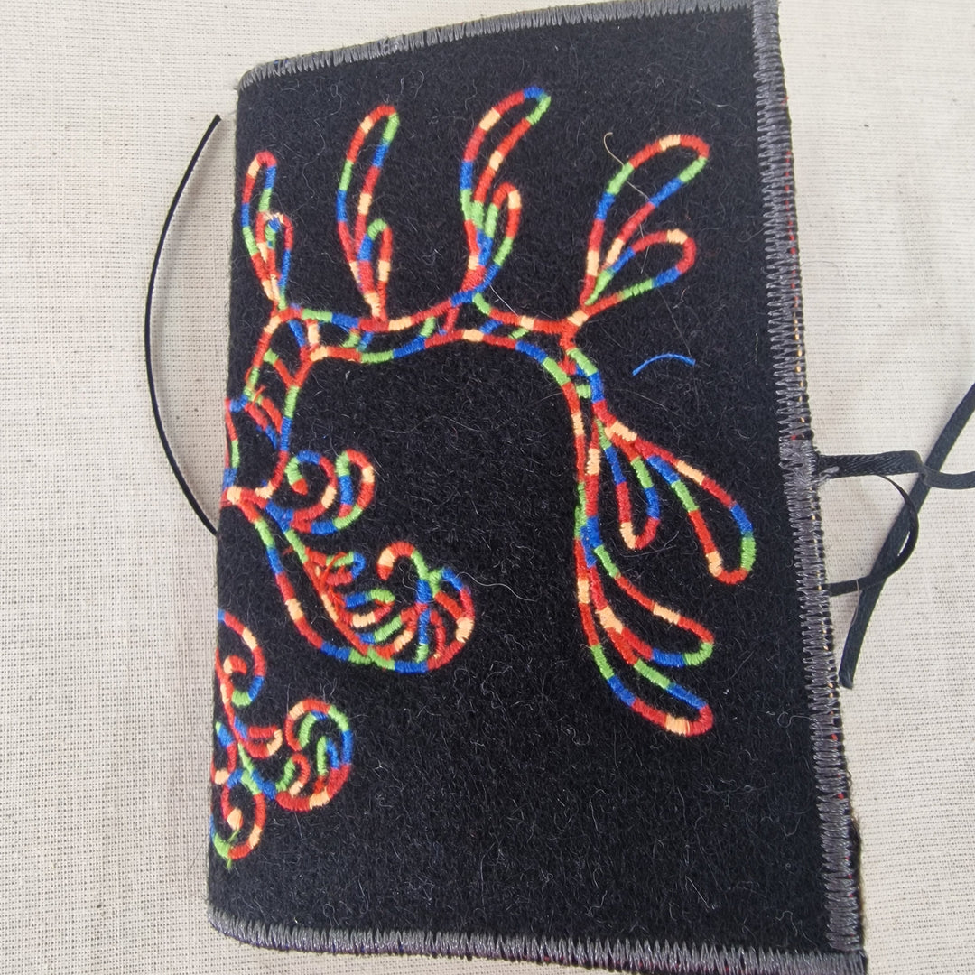 A black felt Needlecase featuring a colourful embroidered seahorse tail design, shown on a neutral beige textured fabric background.