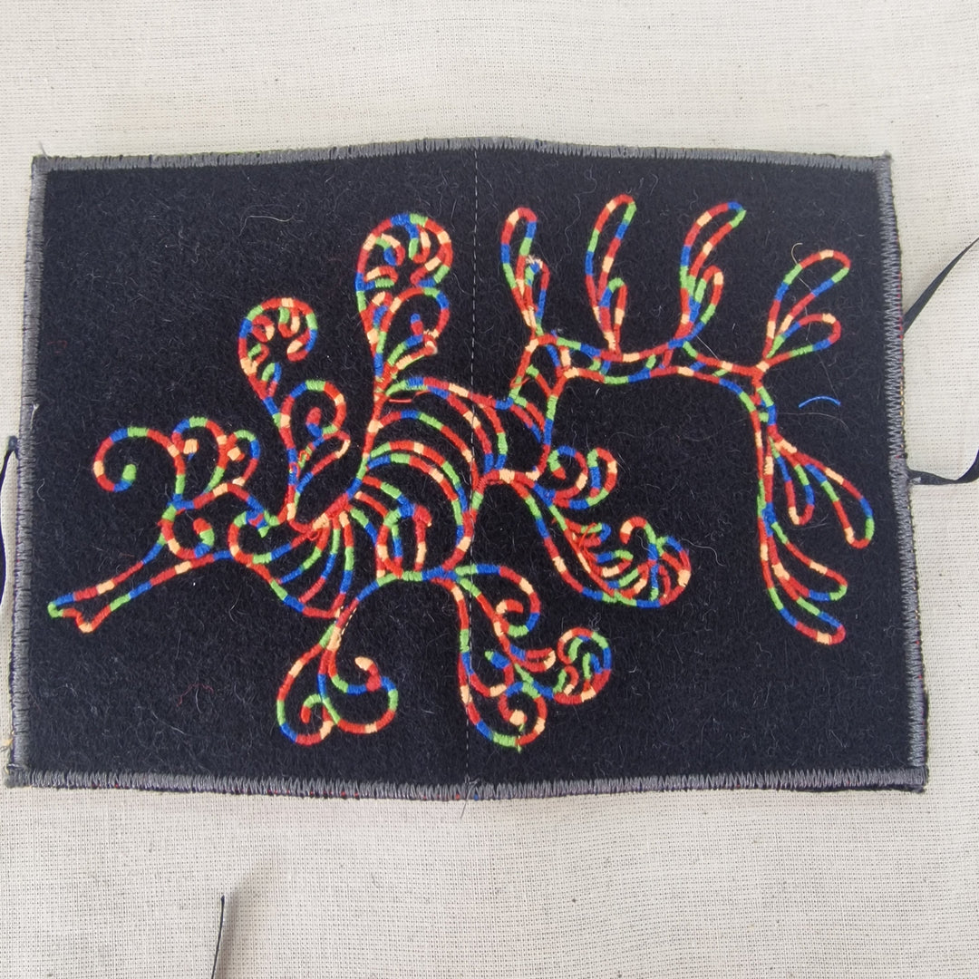 A square black fabric Needlecase featuring a colourful embroidered seahorse design in red, blue, green, and yellow threads, laid flat on a light beige linen background.