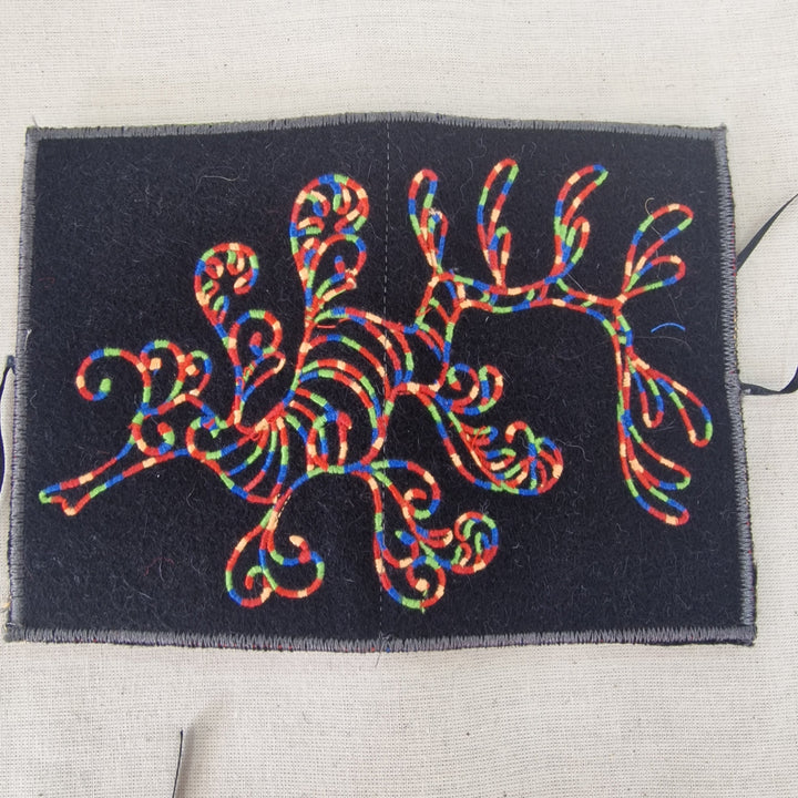 A square black fabric Needlecase featuring a colourful embroidered seahorse design in red, blue, green, and yellow threads, laid flat on a light beige linen background.