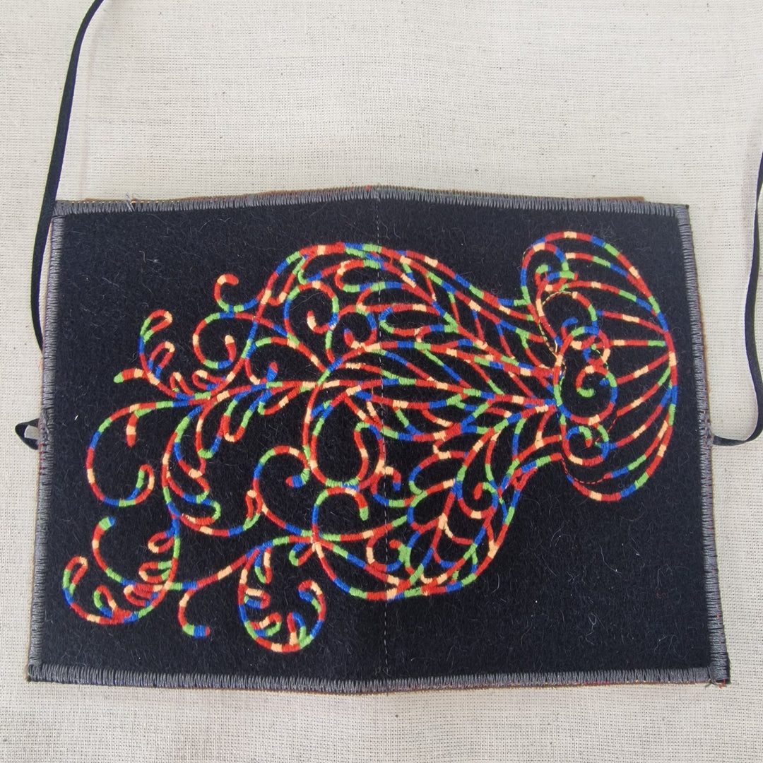 A rectangular black fabric Needlecase with a colourful embroidered scrollwork  Jelly fish design on the front, laid flat on a light beige textured fabric background.