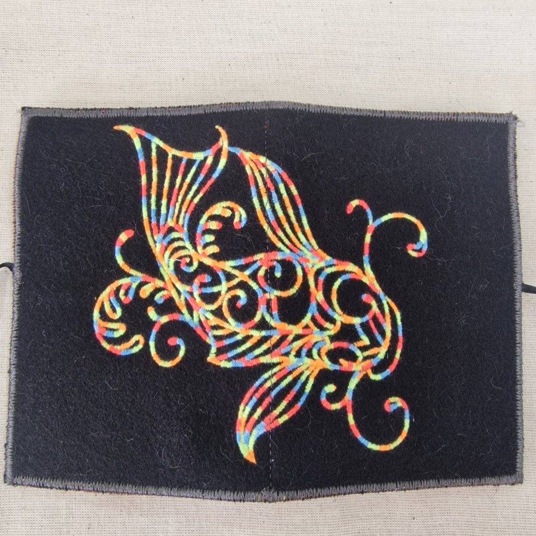 A square black fabric Needlecase featuring a colorful embroidered fish design with swirling, ornate patterns in a rainbow of threads, displayed on a light beige textured fabric background.