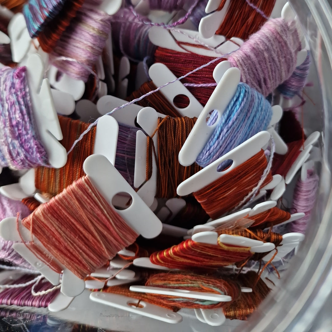 Assorted colorful embroidery threads on white bobbins inside a clear container.